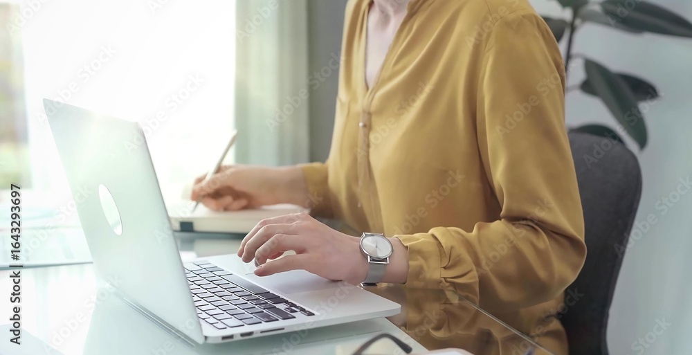 Naklejka premium Unrecognizable professional woman is making notes and working with laptop in modern office. Yellow blouse is passing to adult businesswoman. Business concept