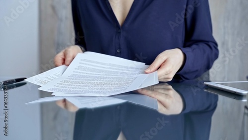 Businessperson woman in dark blue blouse analyzing document at glass desk. Close-up of a professional auditor or lawyer reviewing a lengthy paper report in office setting. Business people concept