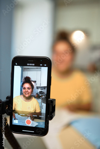 Selective focus om smartphone mounted on tripod recording female blogger in home clothes with hair bun making content or tutorial video for students, tips and advice on effective learning