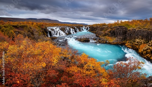 a stunning blue river flows over rocky terrain in iceland creating multiple cascades and surrounded by autumn colored vegetation under a cloudy sky