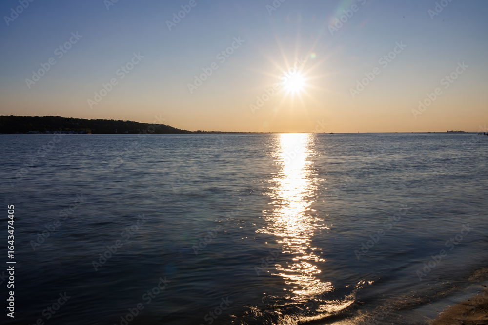 Naklejka premium Golden sunset over calm ocean waves, with shimmering reflections stretching to the horizon, tranquil coastal scenery, and a clear blue sky perfect for travel, nature, and summer landscape themes.