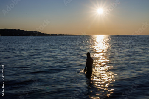 Silhouette of a person standing in calm sea waters during a golden sunset, with shimmering reflections and a tranquil horizon under a warm evening sky.