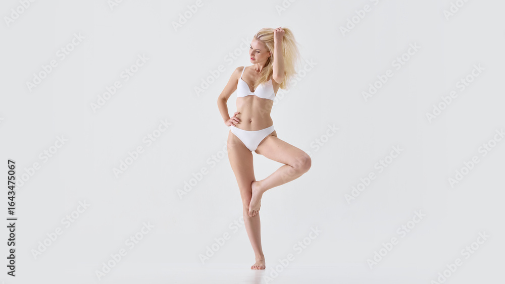 Fototapeta premium Blonde woman in white lingerie posing playfully with hand in hair and confident smile. Concept of fashion catalog, lingerie promotion, feminine charm, elegance and body positivity.