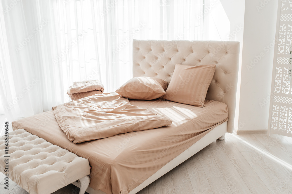 Naklejka premium Cozy beige bed with soft pillows and blanket in bright sunlight
