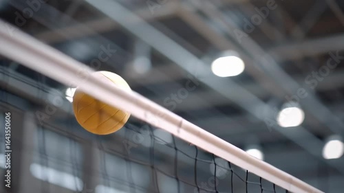 Volleyball ball flying over net in indoor sports championship game with bright lights creating exciting dynamic action atmosphere