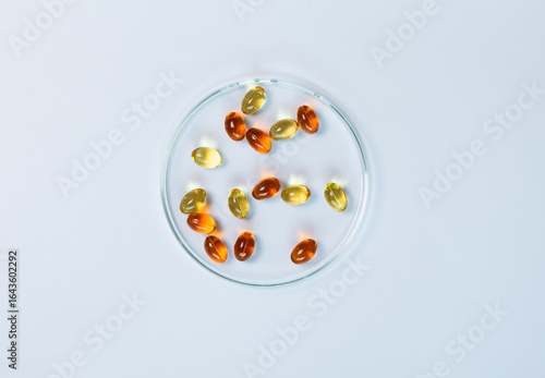 Top-view of yellow and amber supplement capsules in a glass petri dish, isolated on a white background. This shows scientific research, pharmaceutical development for health products like fish oi.
