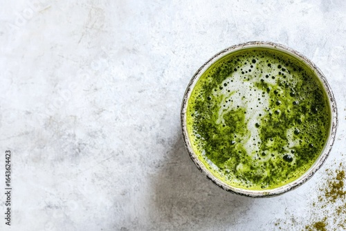Overhead shot of a vibrant green matcha latte in a light-colored bowl, atop a mottled gray surface; a sprinkle of matcha powder is visible nearby.  The latte displays a creamy, frothy texture