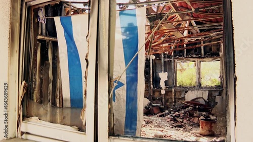 The broken window as the frame, looking inside the completely destroyed and burnt living room, where an Israeli flag is hanged sadly. Kibbutz Nir Oz, southern Israel massacre memorandum.