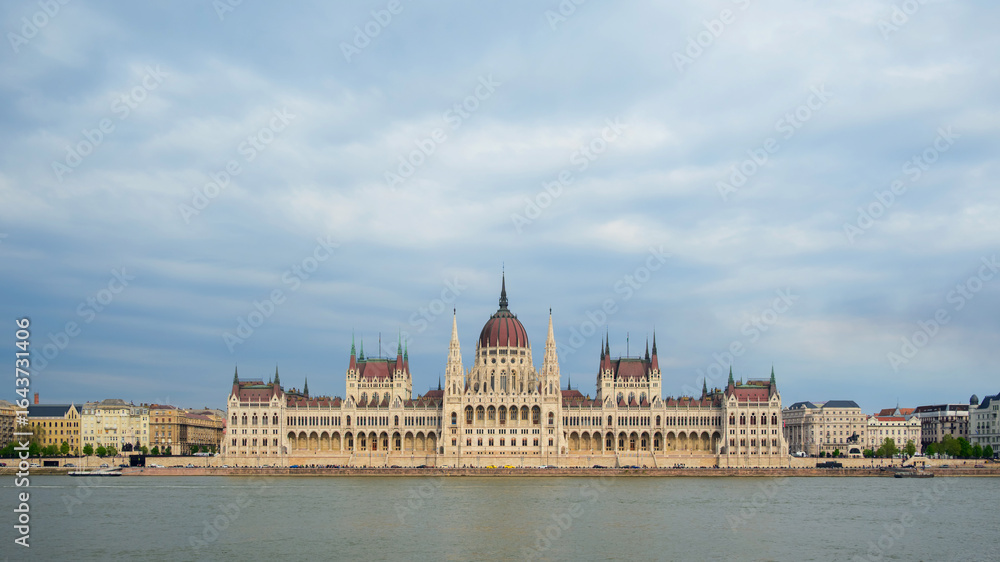 Obraz premium Visitors admire the majestic Hungarian Parliament Building in Budapest, reflecting on the Danube River, showcasing its intricate architecture and historic significance during the day.