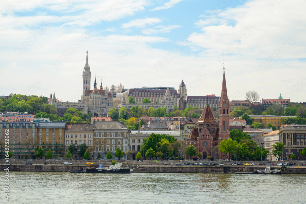 Fototapeta premium Budapest's skyline showcases a blend of historic and contemporary architecture along the Danube, with green hills and vibrant buildings. A bridge connects the urban landscape and nature.