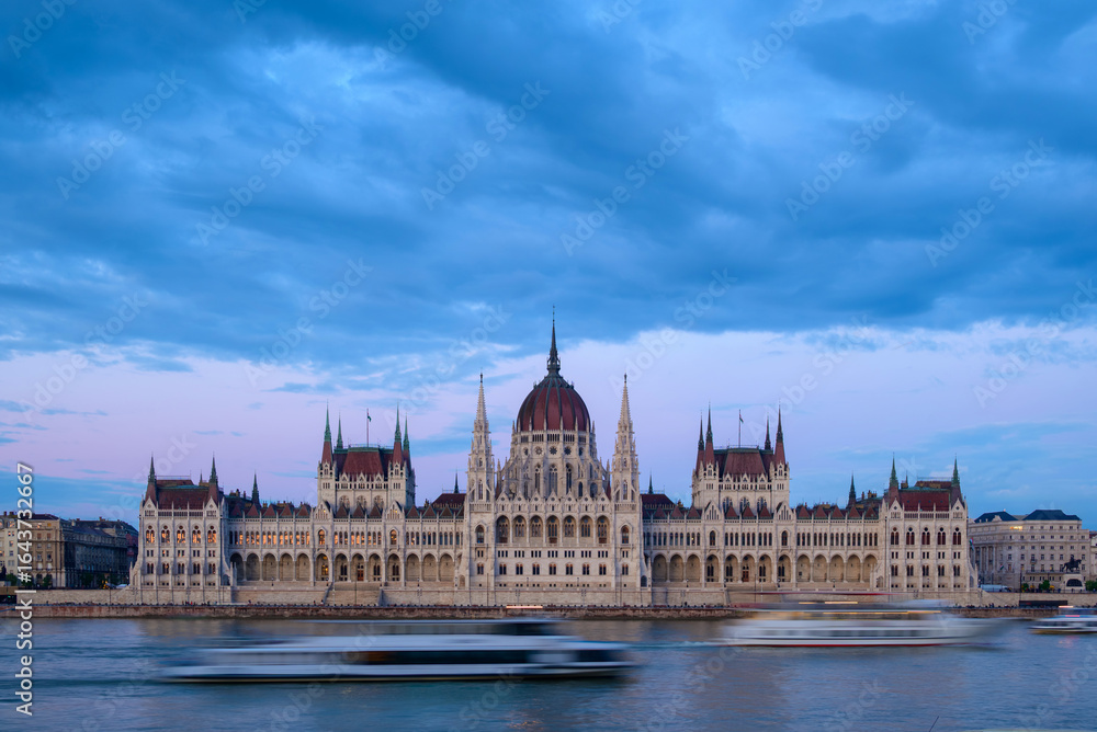 The majestic Hungarian Parliament building stands along the Danube River as the sky transitions to twilight. Boats glide past, reflecting the iconic architecture in the water. The majestic Hungarian Parliament building stands along the Danube River as the sky transitions to twilight. Boats glide past, reflecting the iconic architecture in the water.