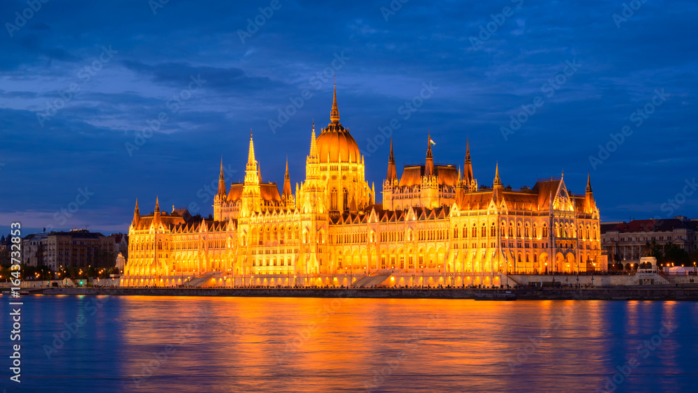 Fototapeta premium The majestic Hungarian Parliament Building stands illuminated against the darkening sky, reflecting beautifully on the Danube River in Budapest during the evening.