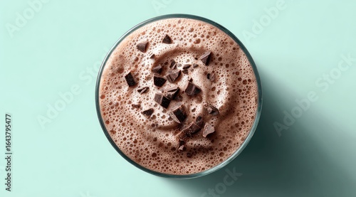 Frothy chocolate milkshake in a clear glass, topped with chocolate shavings, viewed from above against a pale teal background