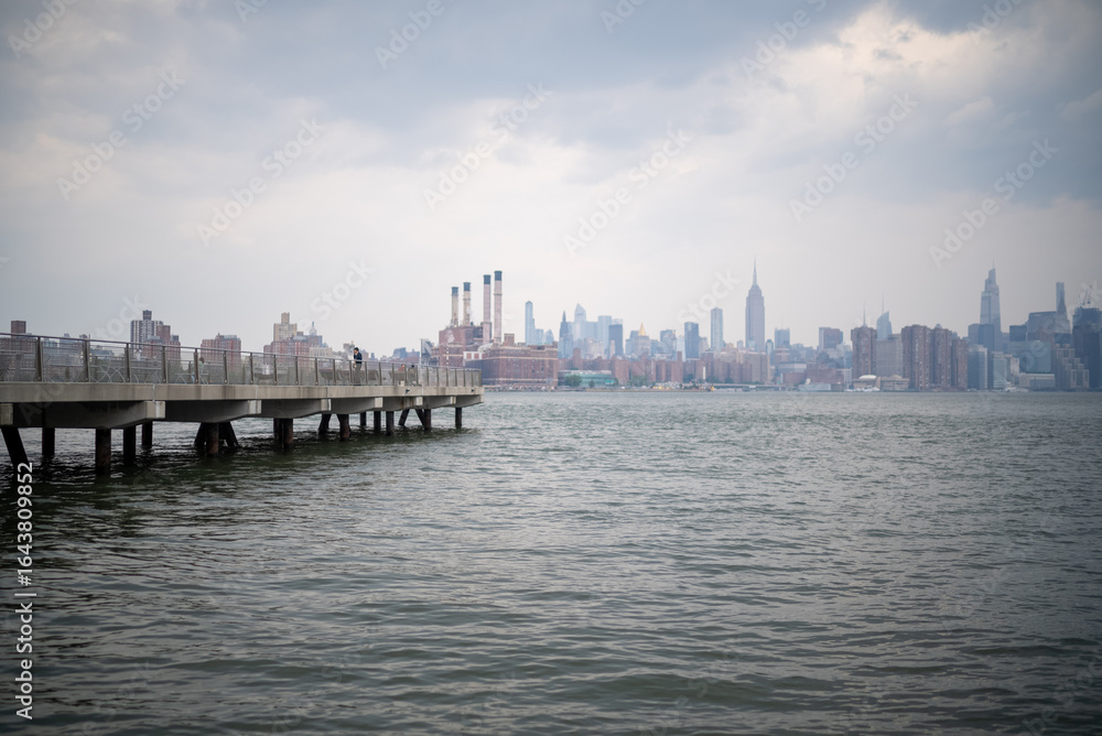 Naklejka premium Pier extends into the water while city skyline looms under cloudy sky in New York during late afternoon