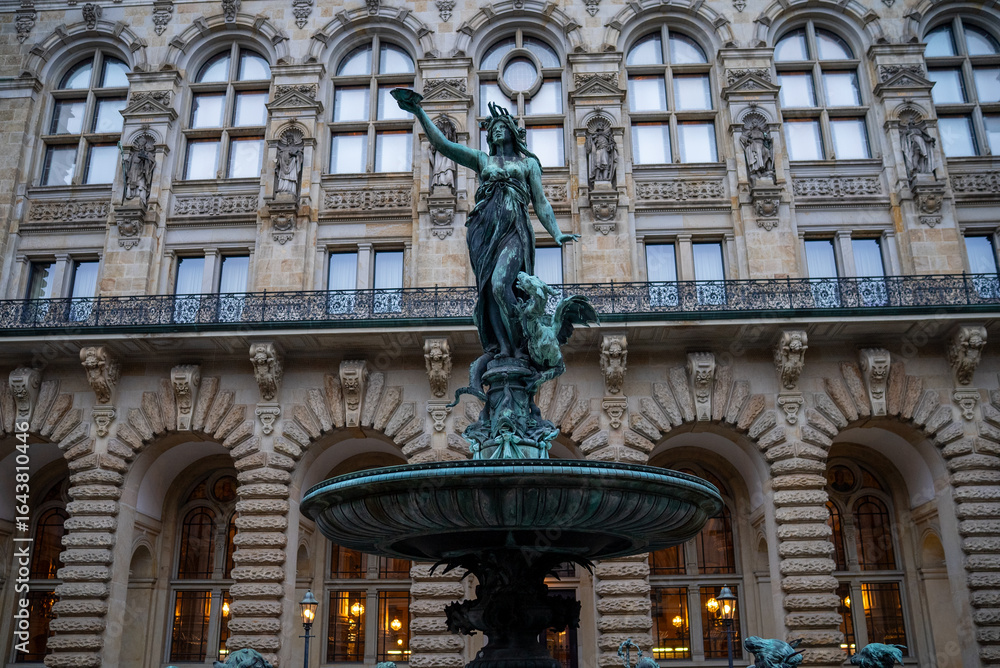 Obraz premium Majestic fountain featuring a woman holding a bowl, set in front of ornate building architecture in an urban plaza during early evening hours