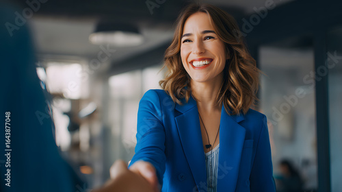 Professional businesswoman in blue blazer smiling and shaking hands with colleague, close-up shot, modern office background with soft natural lighting, confident and friendly expression, cinematic dep