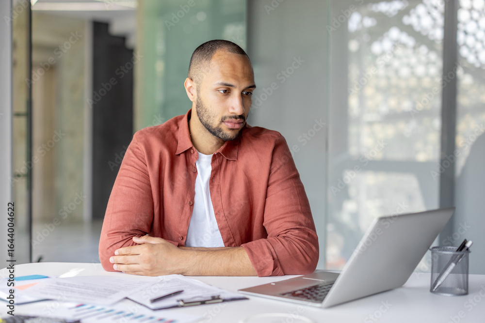 Naklejka premium Focused young man wearing a casual shirt working remotely on his laptop in a modern environment with documents nearby. The setting suggests productivity, professionalism, and a relaxed workspace.