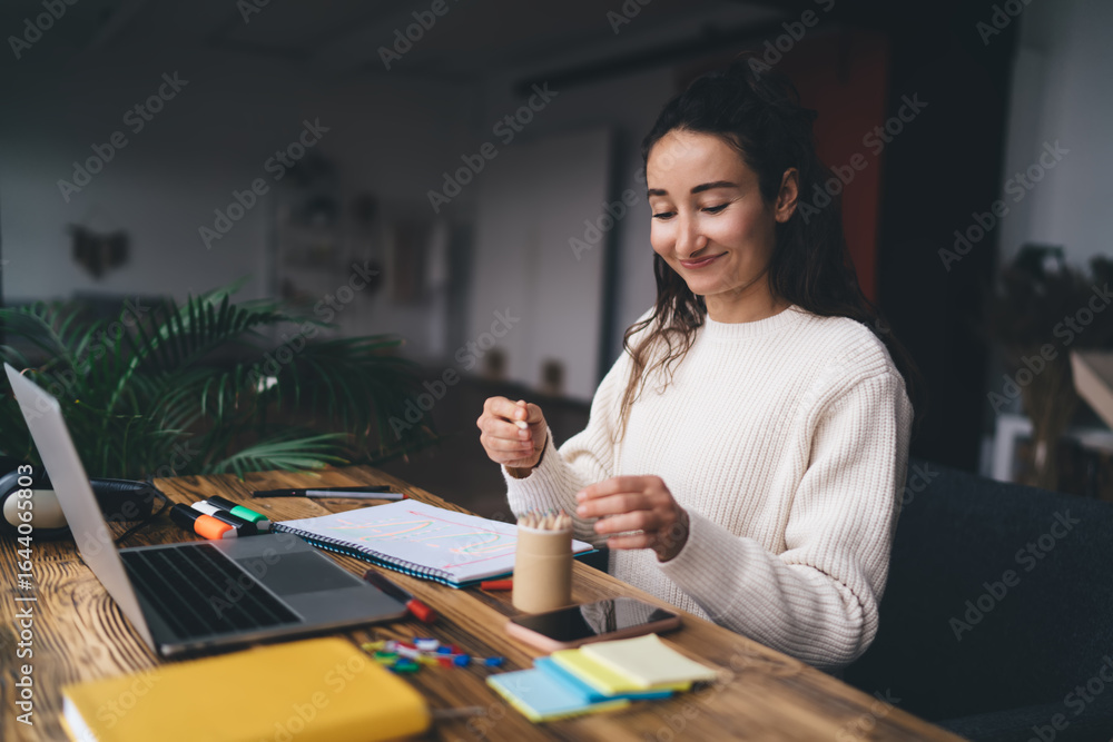 Fototapeta premium Young woman freelancer in cozy sweater choosing pencil for creative planning near laptop, representing inspiration, balance and productivity in a digitally augmented work process.