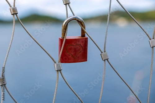 Fotografie A red love lock with a heart symbol locked onto a steel cable fence, representing eternal love and commitment, with a blurred sea background