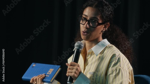 Tilt up shot of young confident biracial female author sitting on stage and speaking into microphone while presenting her new motivational book to unrecognizable audience