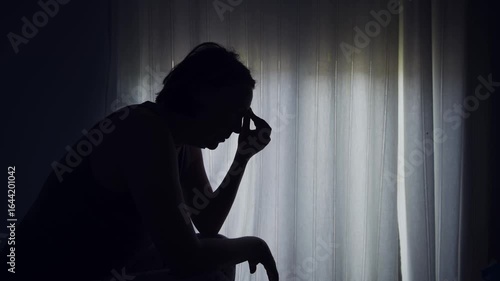 Silhouette of a woman sitting by a window, holding her head in distress, expressing anxiety, sadness, or emotional struggle. 4K.