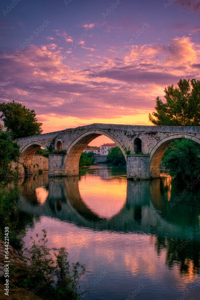 Fototapeta premium A serene sunset illuminates a historic stone bridge, creating stunning reflections in the tranquil river below. Lush greenery frames the scenic view