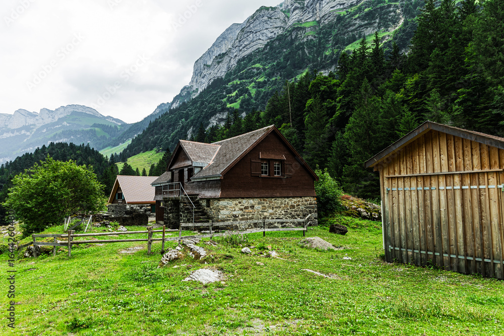 Obraz premium Traditional wooden and stone alpine houses set against a backdrop of lush forest and towering mountains near Seealpsee in the Appenzell Alps, Switzerland.
