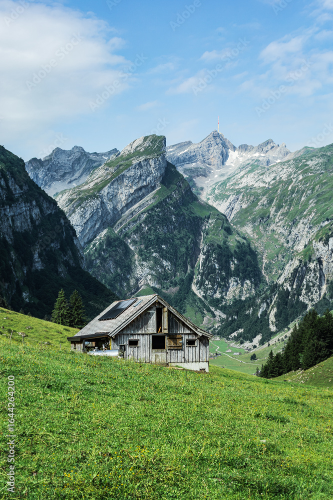 Obraz premium Traditional Swiss alpine house with solar panels on the roof, located on a grassy hillside with dramatic rocky mountain peaks in the background, captured on a clear summer day.