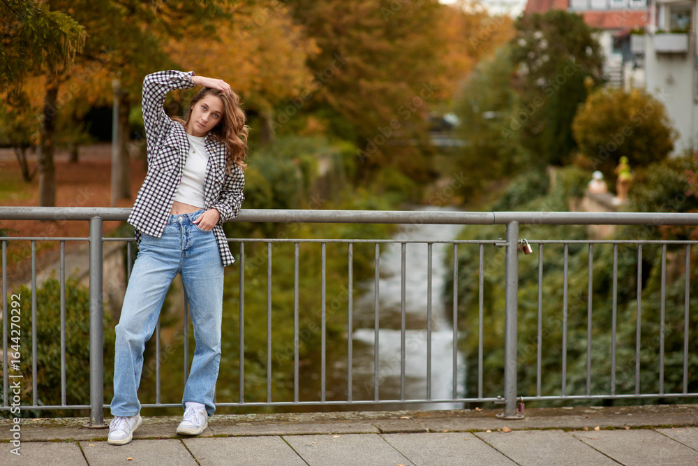 Fototapeta premium Young woman in trendy autumn outfit posing on a European bridge with golden foliage and a historic cityscape in the background.