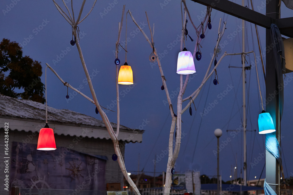Obraz premium Dry tree with hanging lanterns and Turkish amulets of blue glass under window of seaside cafe at dusk. Concept of summer, relaxation, service