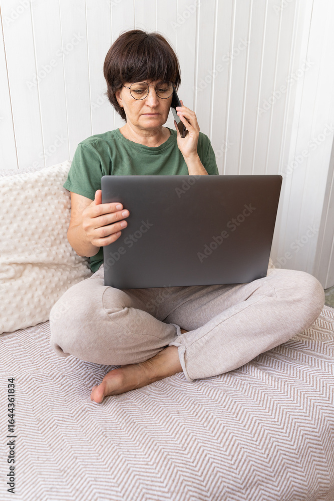 Naklejka premium Caucasian mature woman using laptop and phone on sofa in relaxed home setting