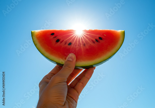 A hand holds a juicy watermelon slice against a vibrant summer sky, backlit by the sun's radiant glow.