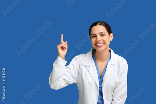 Smiling female doctor in white lab coat pointing upward with index finger, me...