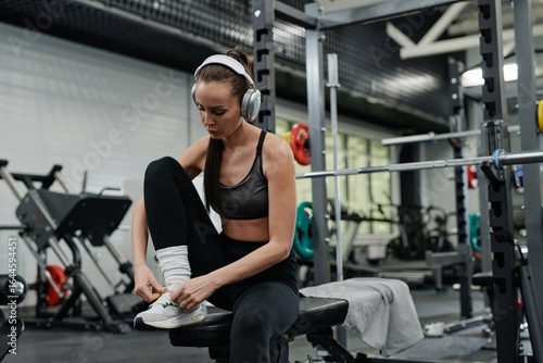 Young woman wearing headphones spending time in gym sitting on bench lacing sneakers, copy space