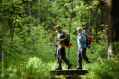 Two senior Caucasian people hiking through lush green forest, carrying backpacks and walking across small wooden bridge, enjoying outdoor adventure and exploring nature together