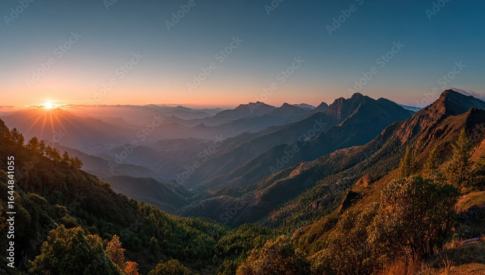Obraz premium Panoramic sunset view over a vast mountain range, layers of hazy hills fading into a clear sky, sun's rays casting a golden glow on the landscape