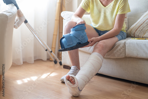 Quadro em tela Woman sitting on sofa with plastered leg, holding a medical boot, recovering fro