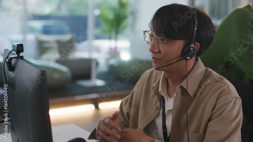 Focused young asian male operator provides diligent customer support from modern call center, professional representative wearing headset assists client during call