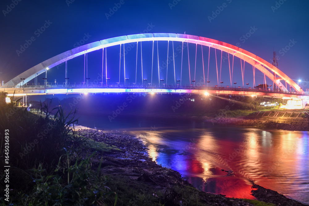 Obraz premium A beautiful bridge with colorful lights stretches over a calm river at night.