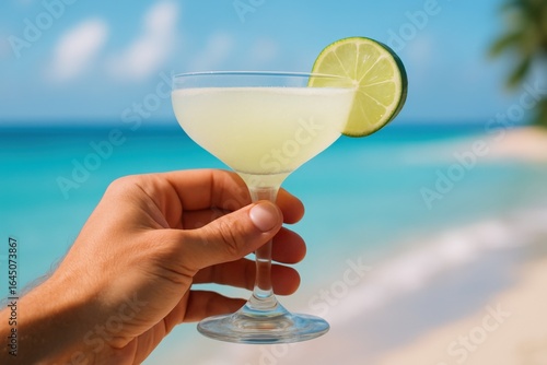 Fototapeta Naklejka Na Ścianę i Meble -  Hand holding cocktail glass with lime on sunny beach