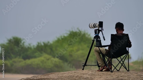 A wildlife photographer and surf videographer sits on coastal rocks beside a tripod-mounted telephoto camera. He waits quietly while filming surfers and scanning the shoreline for wildlife activity.