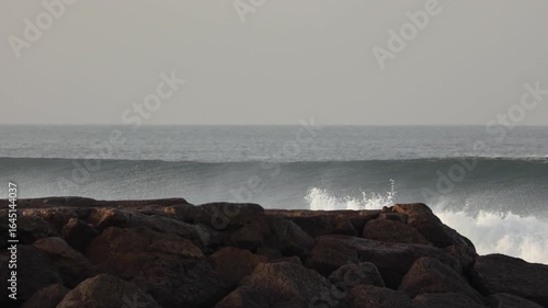 Slow motion pan starting with breaking waves, revealing man in pink sweater on jetty enjoying the view. Cold winter morning, captured in 4K.