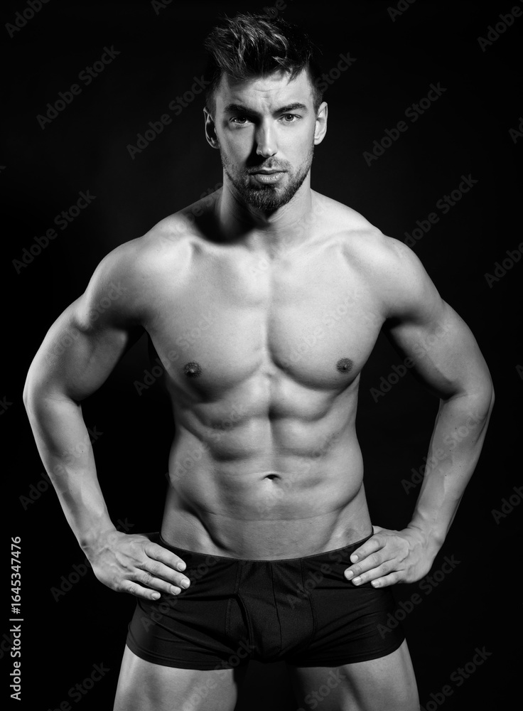 Obraz premium Handsome bodybuilding man posing shirtless on black background flexing muscles and six pack after a workout