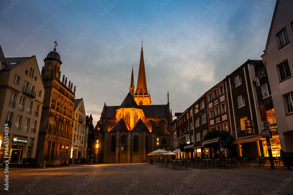 Obraz premium Wesel, Germany – August 6, 2025: Illuminated Willibrordi Cathedral and historic market square at dusk, with surrounding buildings and outdoor cafes.