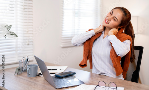 A tired young woman stretches her neck and shoulders to relieve stress and tension while working at her home office desk.