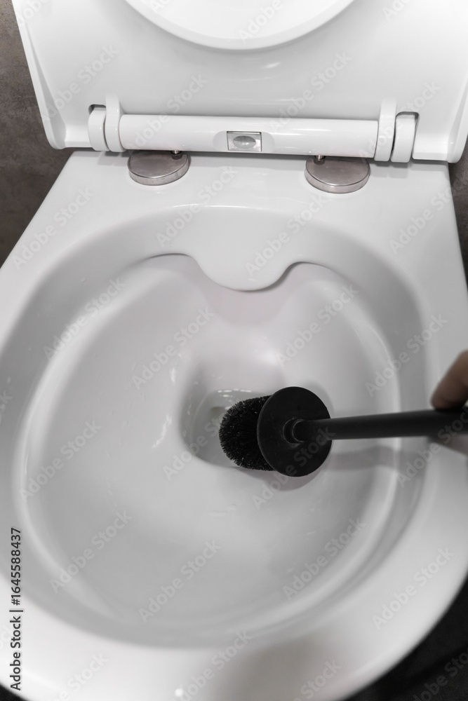 Fototapeta premium Black toilet brush hovering above an open, pristine white toilet bowl, highlighting the importance of bathroom hygiene and promoting a sense of cleanliness and freshness. Vertical photo