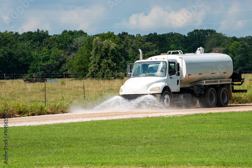 Truck with large tank and powerful pump moves along dirt road as spray nozzels on the front distributes water over the entire road for road maintenance and construction.