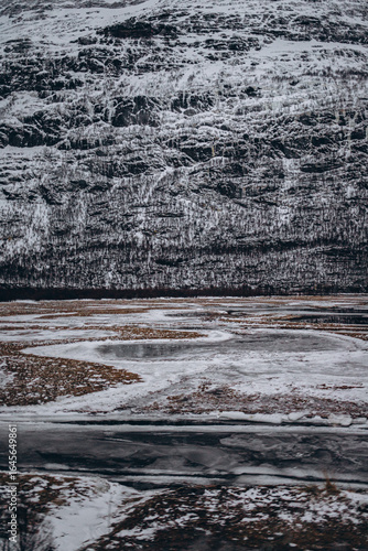 A detailed view of a frozen, winding river or stream in a winter landscape with snow and patches of brown grass, set against a rugged, snow-dusted mountain.