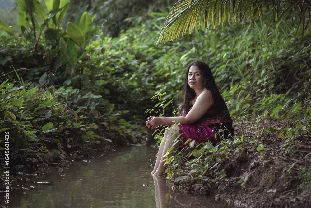 Fototapeta premium Serene Woman Sitting by the Stream Surrounded by Lush Greenery, Enjoying Nature's Beauty in a Tropical Landscape