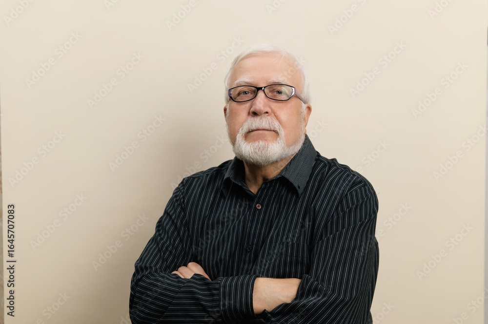 Obraz premium Elderly man with glasses stands confidently against a plain background during a casual indoor portrait session